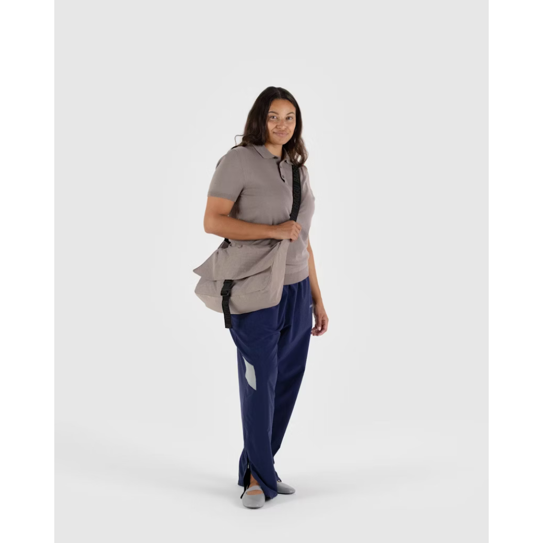 Woman carrying a beige colored messenger bag featuring a long black strap and black buckle closure.