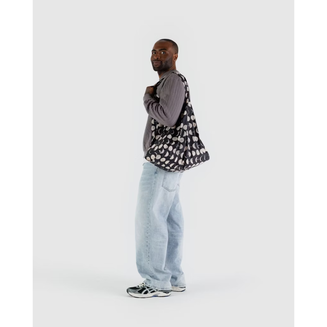 Man carrying a large black reusable tote featuring an all over pattern of white moons in different phases.