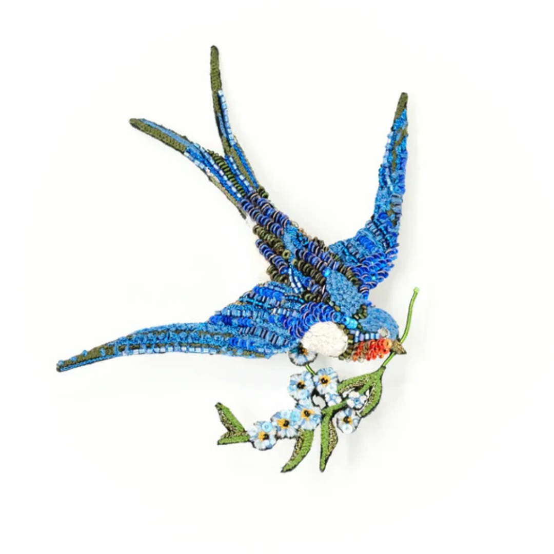 Large hand embroidered and beaded swallow brooch. Bird is blue with a white chest and red chin and carries a branch with flowers in it's beak. The bird is covered in blue beads and micro sequins with white and red sequins on it's neck and chest. The flowers are beaded in delicate light blue and golden yellow with green embroidered leaves and branches.