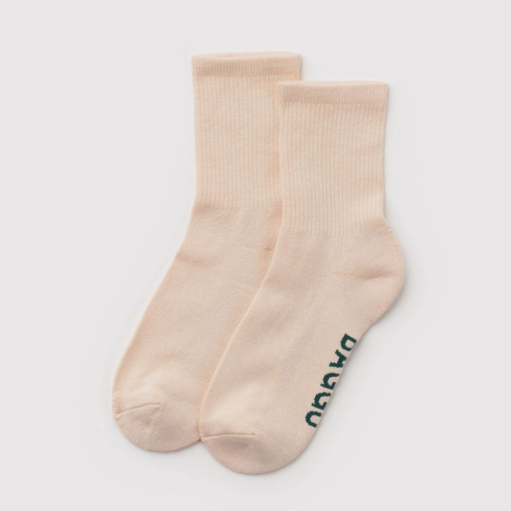 A pair of light peach-beige crew socks with the brand name 'BAGGU' printed in dark teal on the sole of the sock
