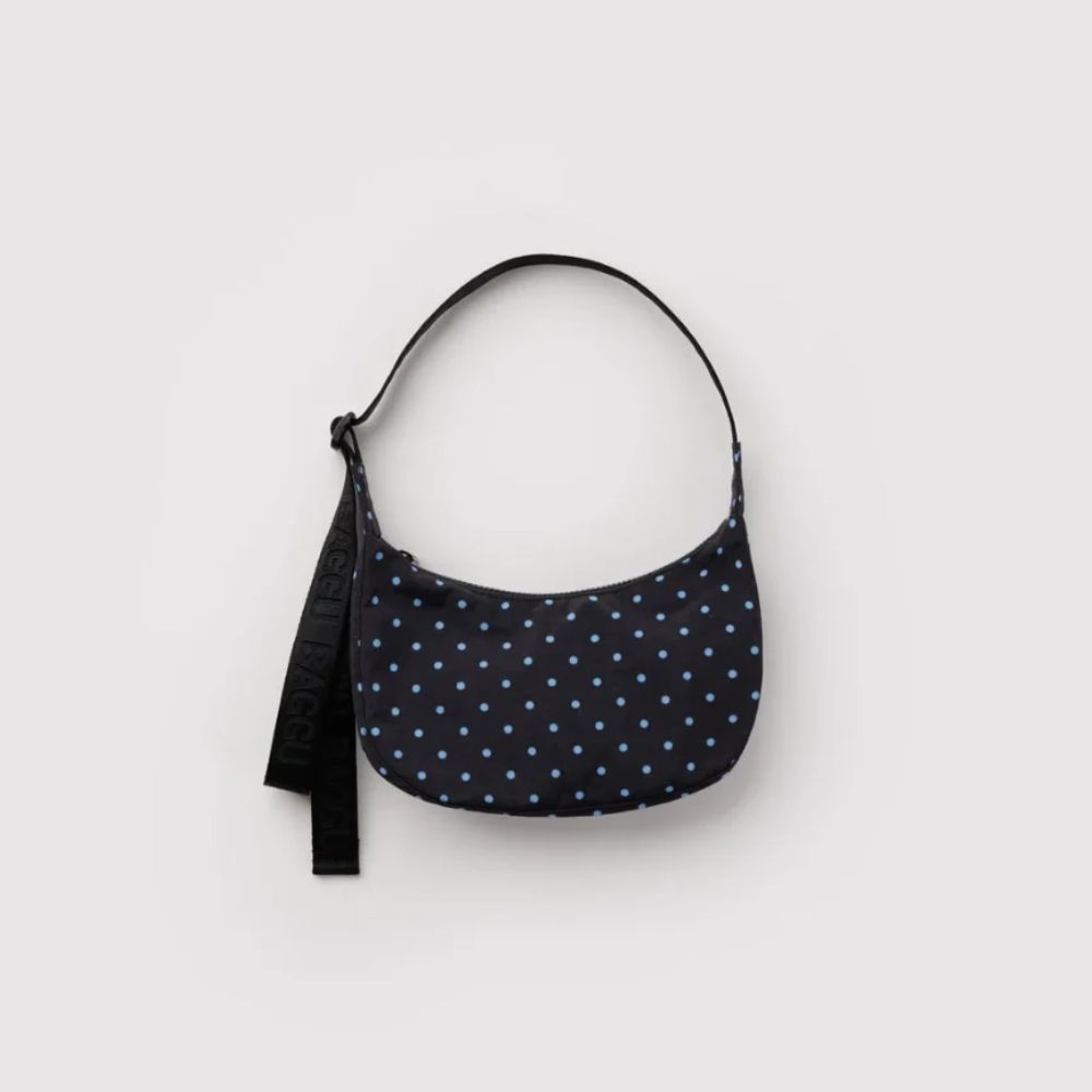 A crescent-shaped shoulder bag with a navy blue base and light blue polka dots, featuring a black adjustable strap, shown against a plain light gray background.