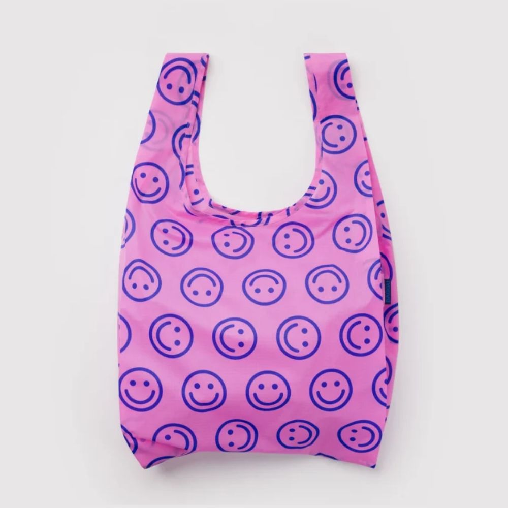 A reusable plastic bag with a pink background and a repeating pattern of dark purple smiley faces, shown against a plain light gray background.