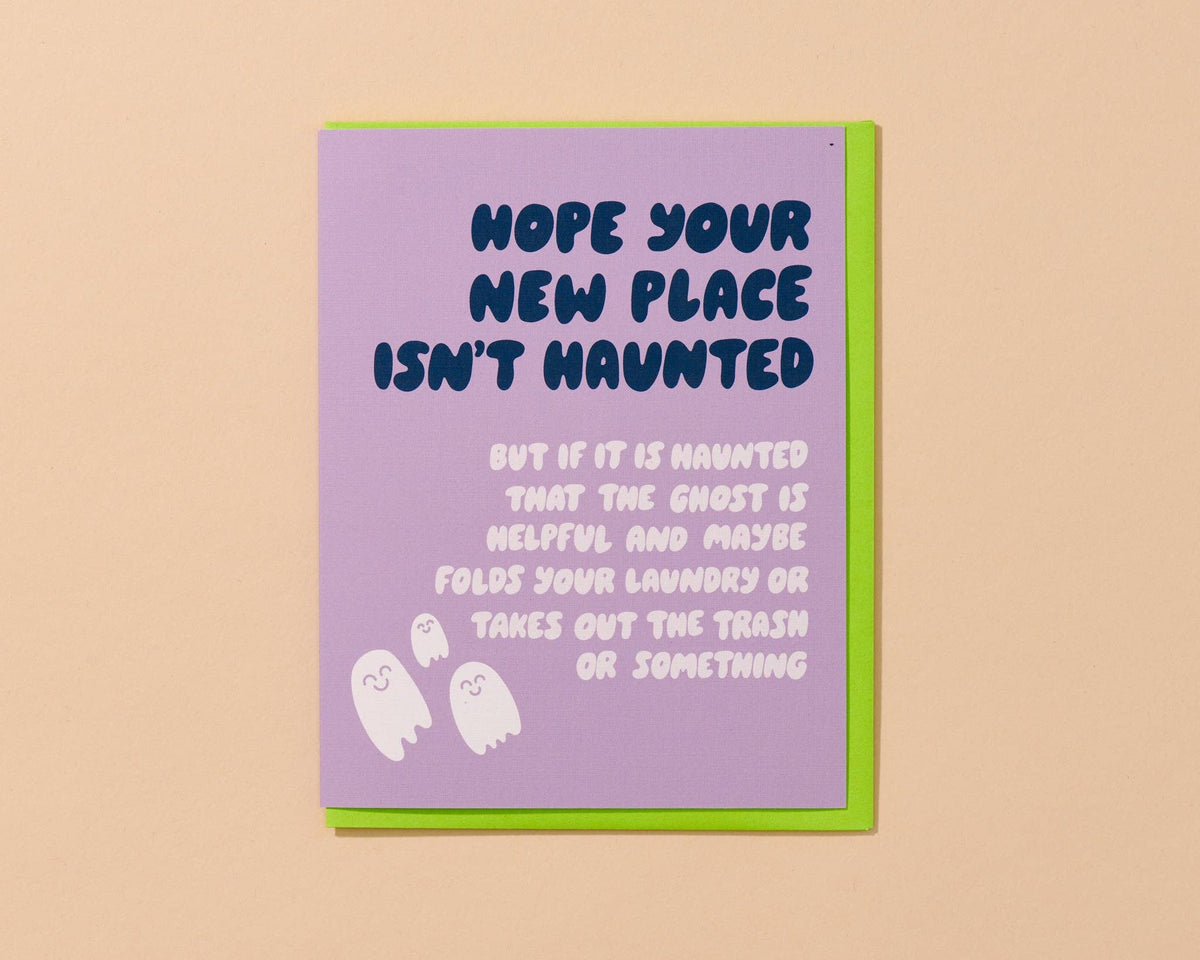 A greeting card with a lavender front and a bright green envelope, shown standing against a warm beige background. The card reads, 'HOPE YOUR NEW PLACE ISN'T HAUNTED BUT IF IT IS HAUNTED THAT THE GHOST IS HELPFUL AND MAYBE FOLDS YOUR LAUNDRY OR TAKES OUT THE TRASH OR SOMETHING,' with small white ghost illustrations at the bottom.