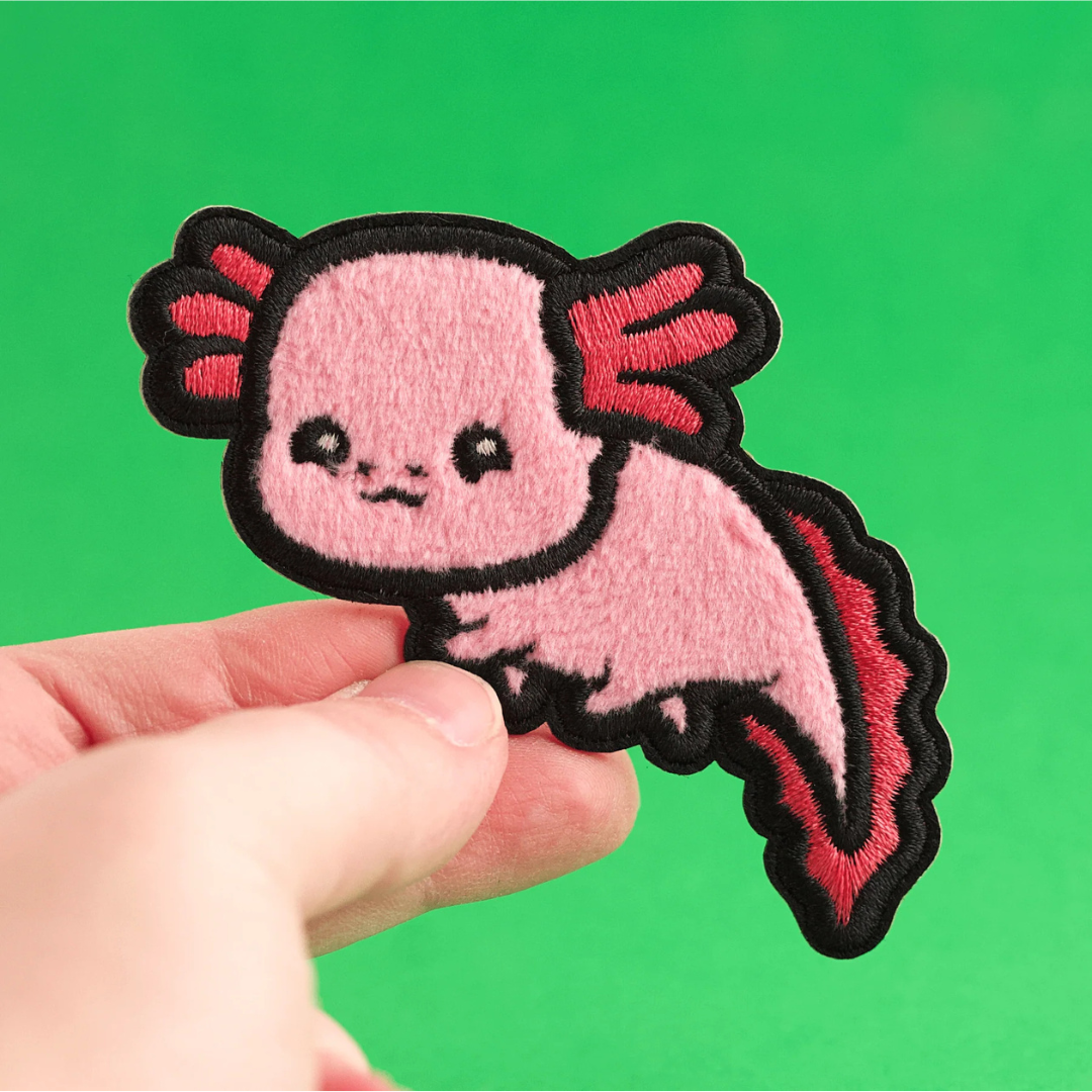 Hand holding a black outlined fuzzy axolotl patch featuring two shades of pink, against a green background.