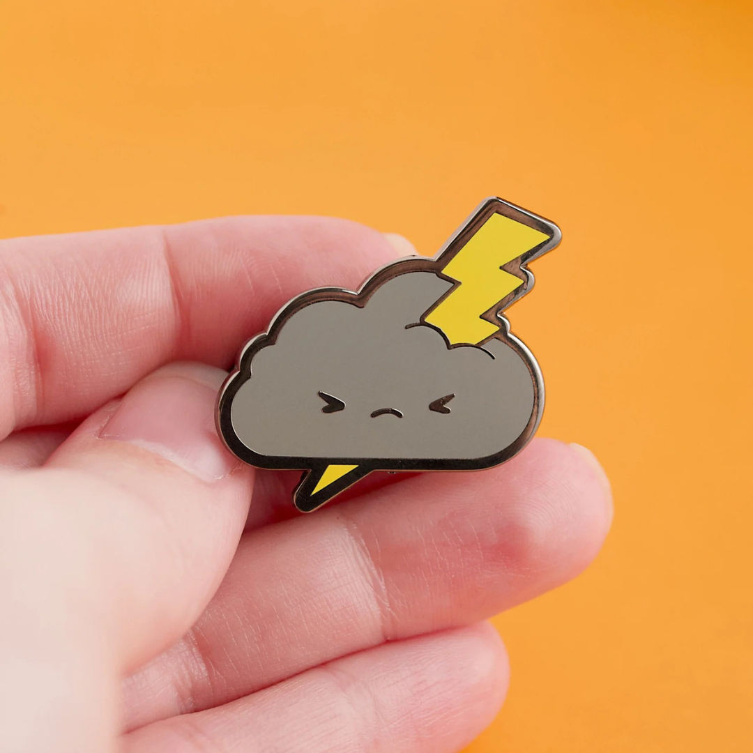 Hand holding a gray cloud pin featuring a grumpy face on it with a yellow lightning bolt through it. All against a golden mustard colored background.