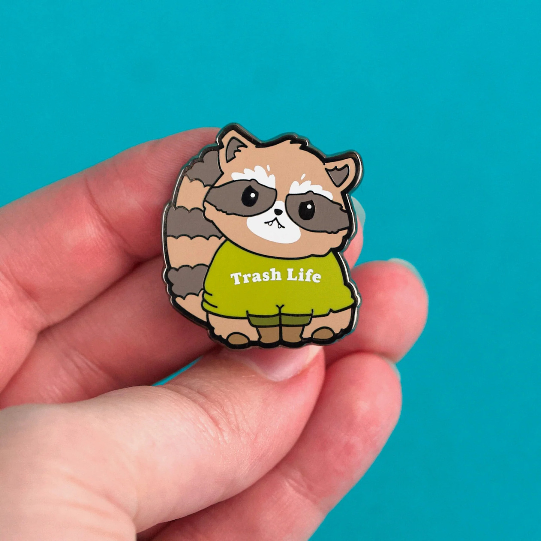 Hand holding a brown and beige raccoon pin wearing a green sweater that reads "Trash Life" in white font. Against a teal background.