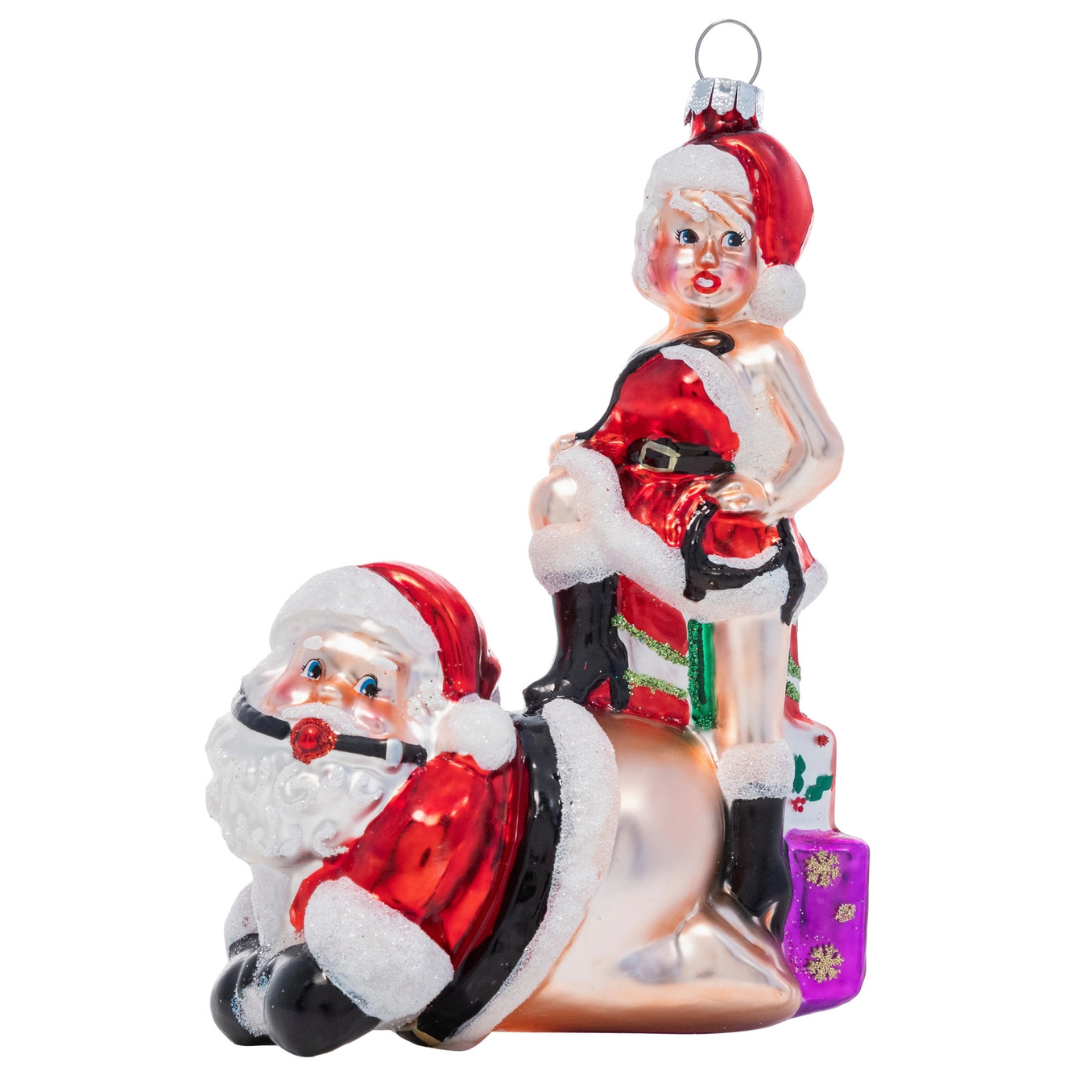 Christmas ornament features a naughty Mrs. Claus standing behind Santa with one leg atop of his back.