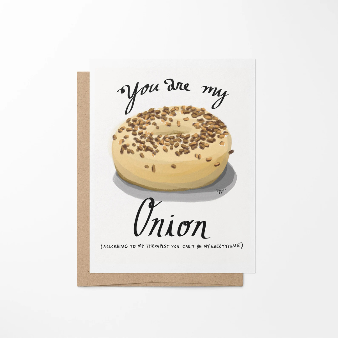 White greeting card featuring a beige and brown onion bagel front and center surrounded by the words "you are my onion (according to my therapist you can't be my everything)" in black lettering.