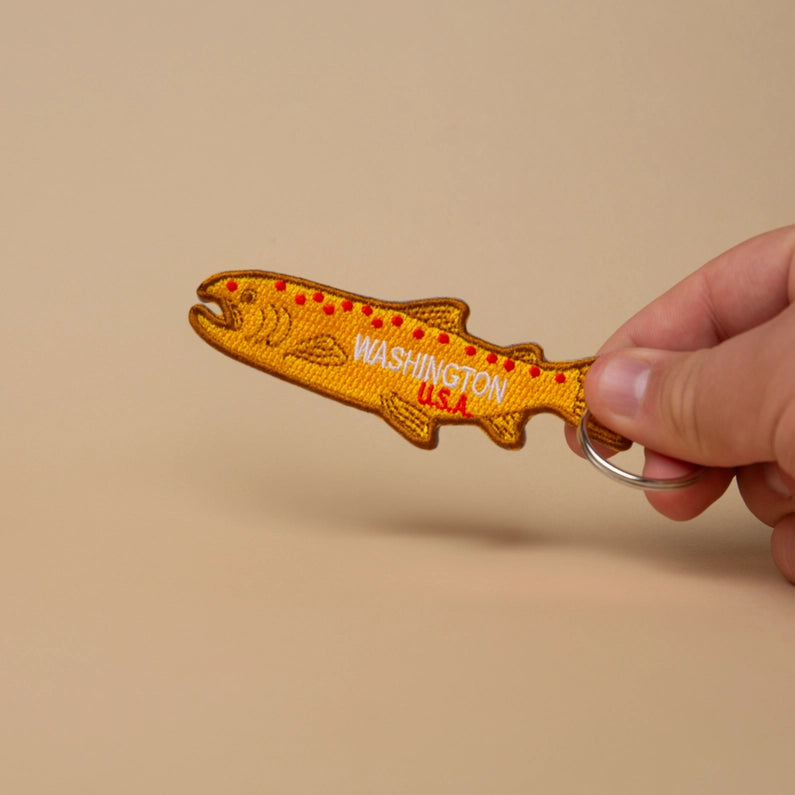 Hand holding a fish-shaped keychain with 'Washington U.S.A' text on a beige background