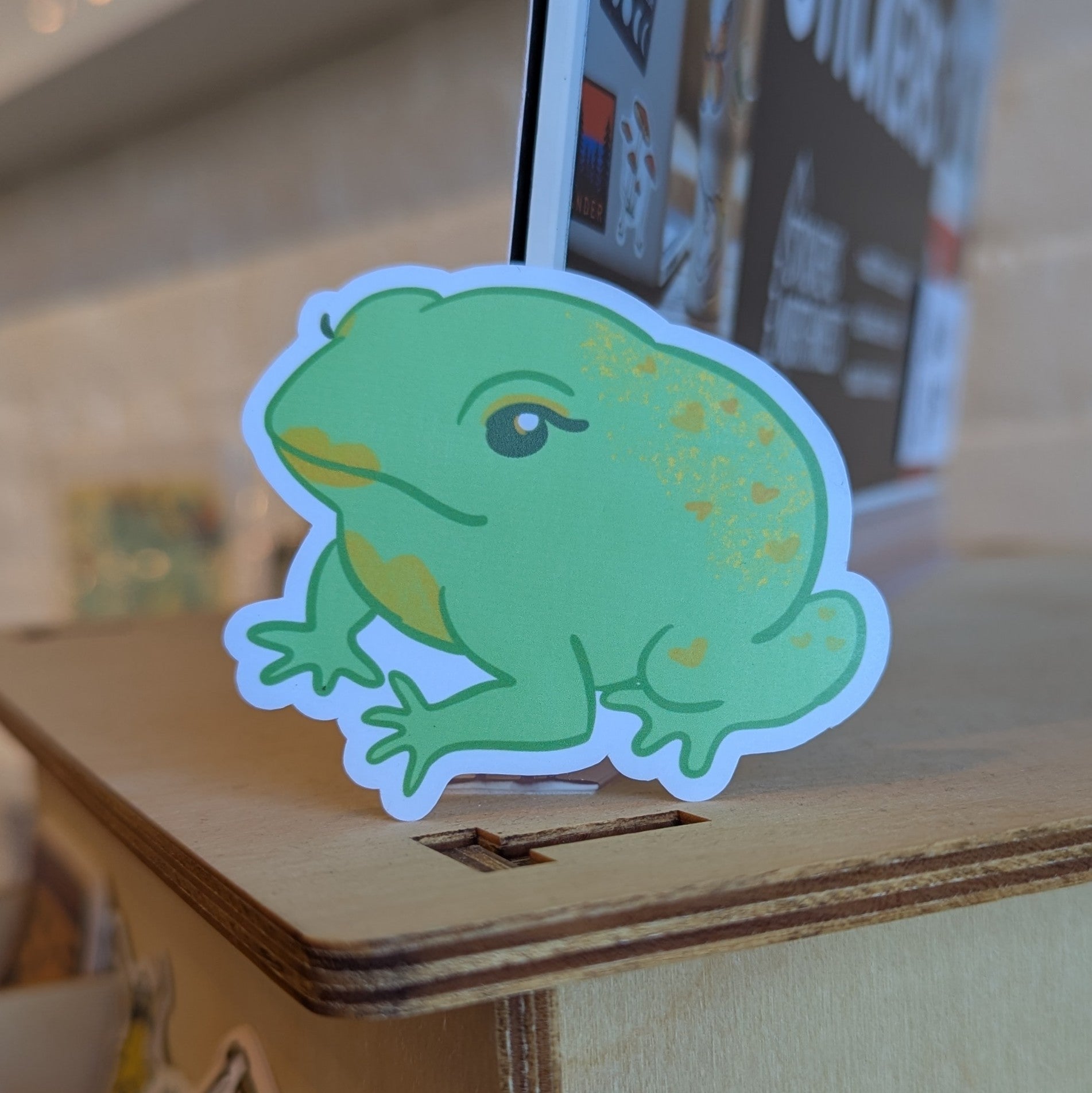 Green frog sticker with gold lipstick and eye makeup. Frog has gold filigree pattern on their back