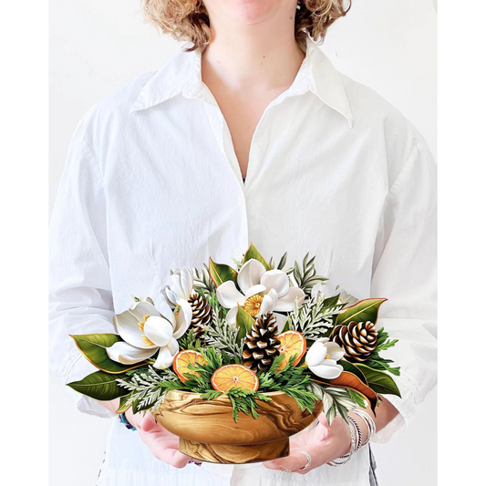 Woman in white holding white winter magnolia bouquet pop-up greeting card. Bouquet filled with white magnolias, pinecones, citrus fruit and winter greenery.