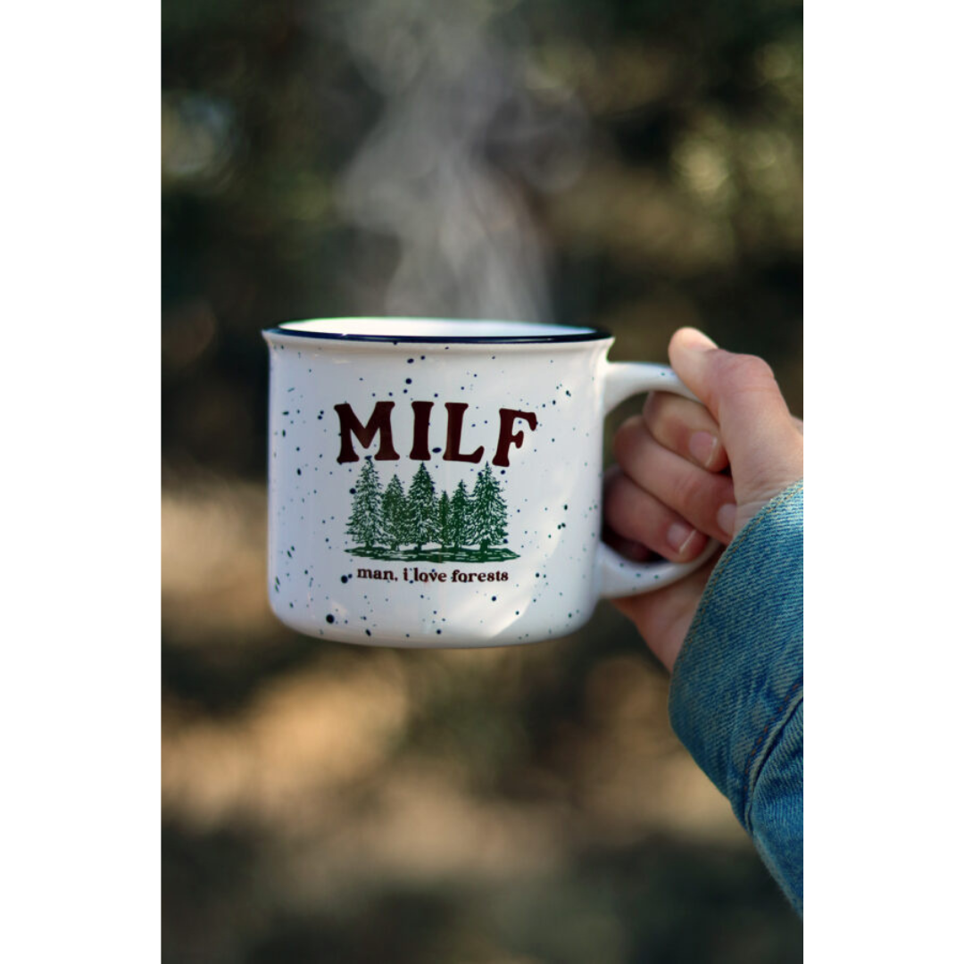 Woman holding off-white speckled ceramic mug with green forestry printed on it featuring brown lettering that reads "MILF man I love forests".