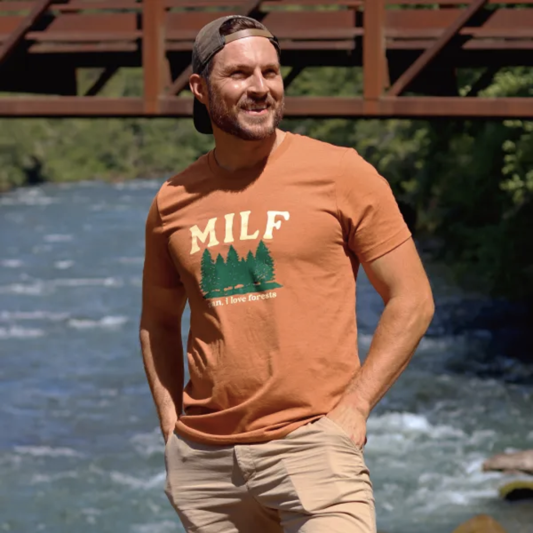 Man standing by a river wearing the burnt orange MILF shirt. Shirt features a forest and says "MILF/man, i love forests"