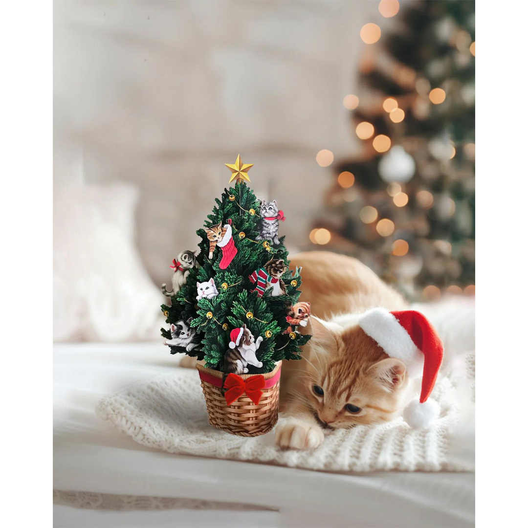 Mini christmas tree pop-up card next to orange kitten wearing a red santa hat, all cozy on a fuzzy white blanket.