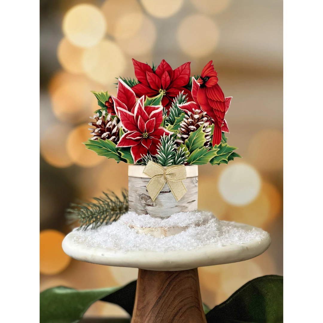 Holiday pop-up greeting card featuring a bright red poinsettia bouquet, pinecones and a cardinal. Tied together with a birch toned bow and sitting on top of a snowy marble tabletop.