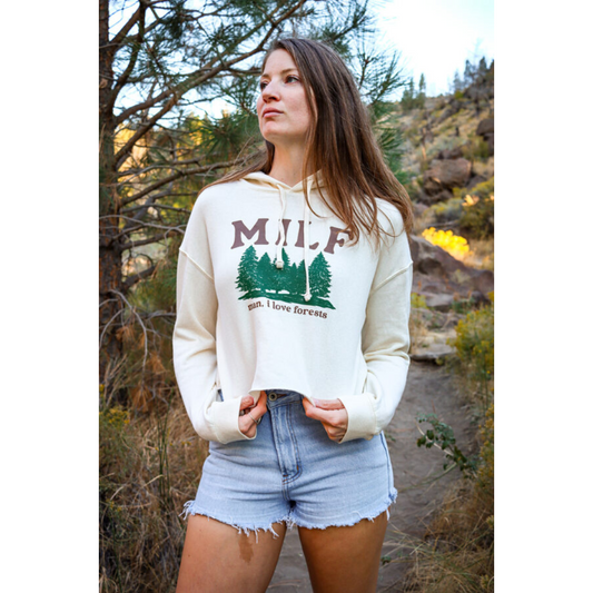 Brunette woman on hiking trail wearing denim shorts and off-white hoodie with green forestry printed on it also featuring brown lettering that reads "MILF man I love forests".