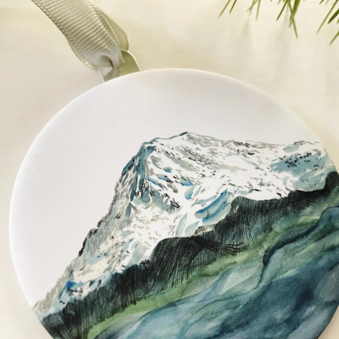 Round metal ornament featuring a watercolor painted picture of Mount Rainier in shades of blue and green. Strung by a grosgrain ribbon.