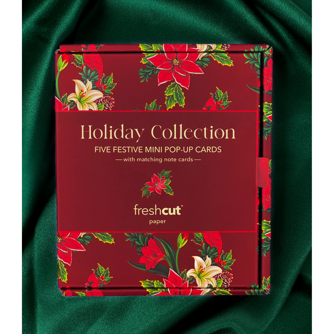 Red holiday box featuring poinsettias with the words "Holiday Collection: Five Festive Mini Pop-Up Cards", sitting on top of emerald green satin.