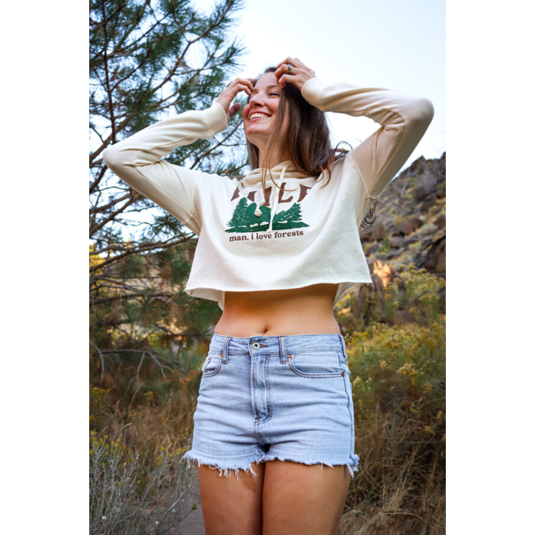 Brunette woman laughing on hiking trail wearing denim shorts and off-white hoodie with green forestry printed on it also featuring brown lettering that reads "MILF man I love forests".