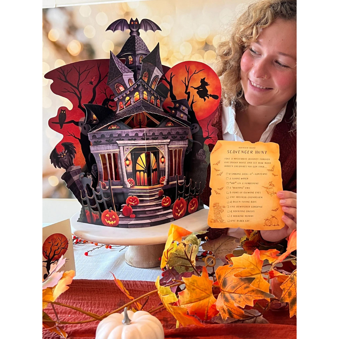 Haunted house pop-up card sitting on a table with woman eyeing in while holding included scavenger hunt directions.