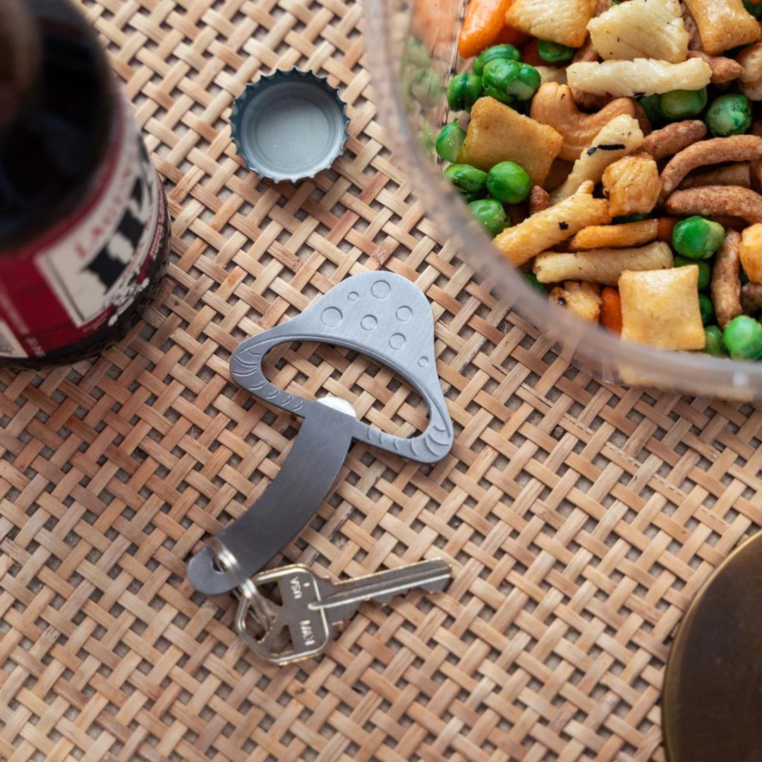 Silver mushroom shaped bottle opener on silver key ring sitting on a rattan tabletop with a beer bottle and bowl of festive snack mix.