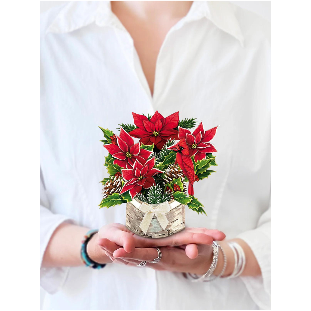 Woman in white holding a holiday pop-up greeting card featuring a bright red poinsettia bouquet, pinecones and a cardinal in the palm of her hands,