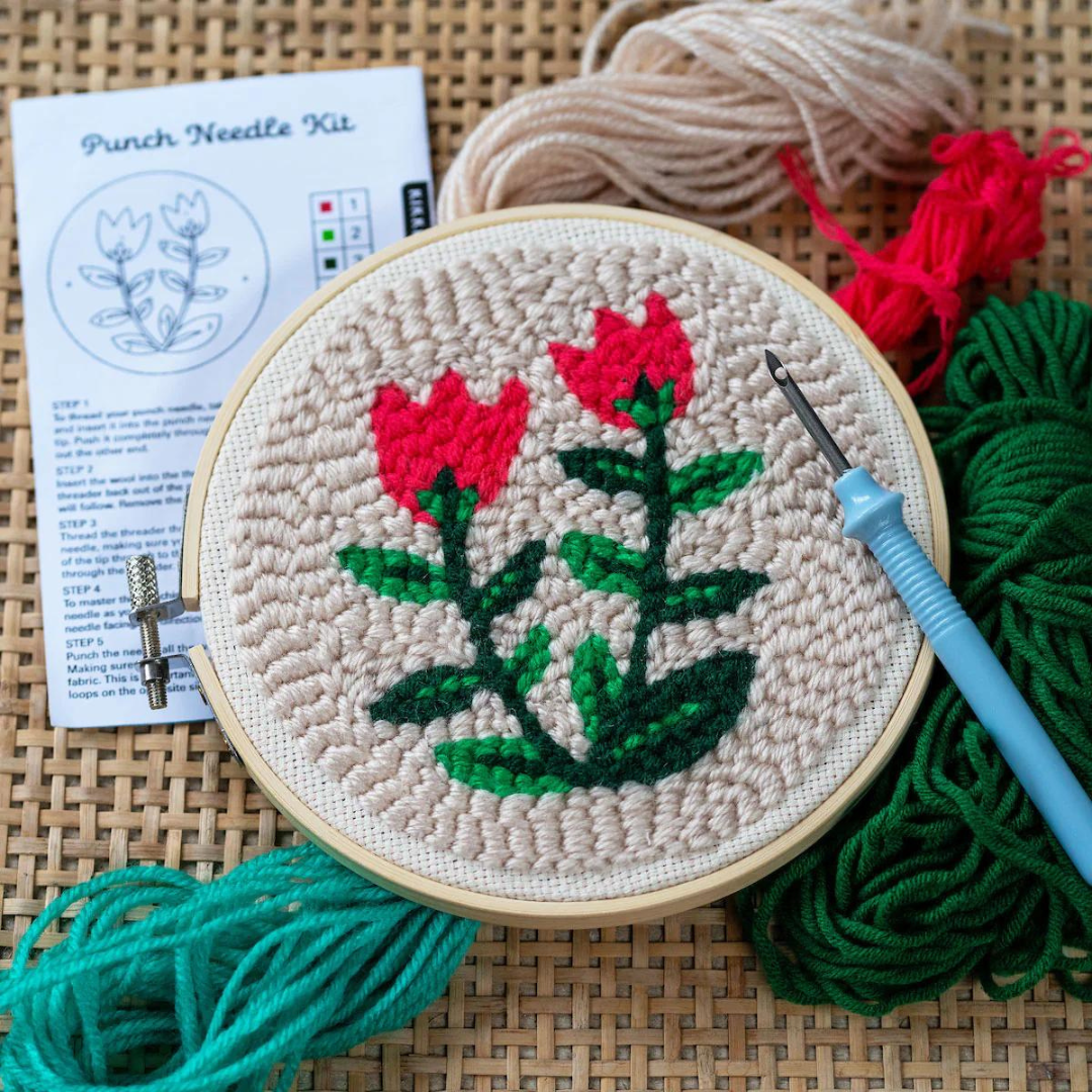 Completed punch needle kit featuring a beige background with red tulips and multi-toned green stems. Shown with extra beige, red and green threads, needle and instructions all sitting on a rattan surface.