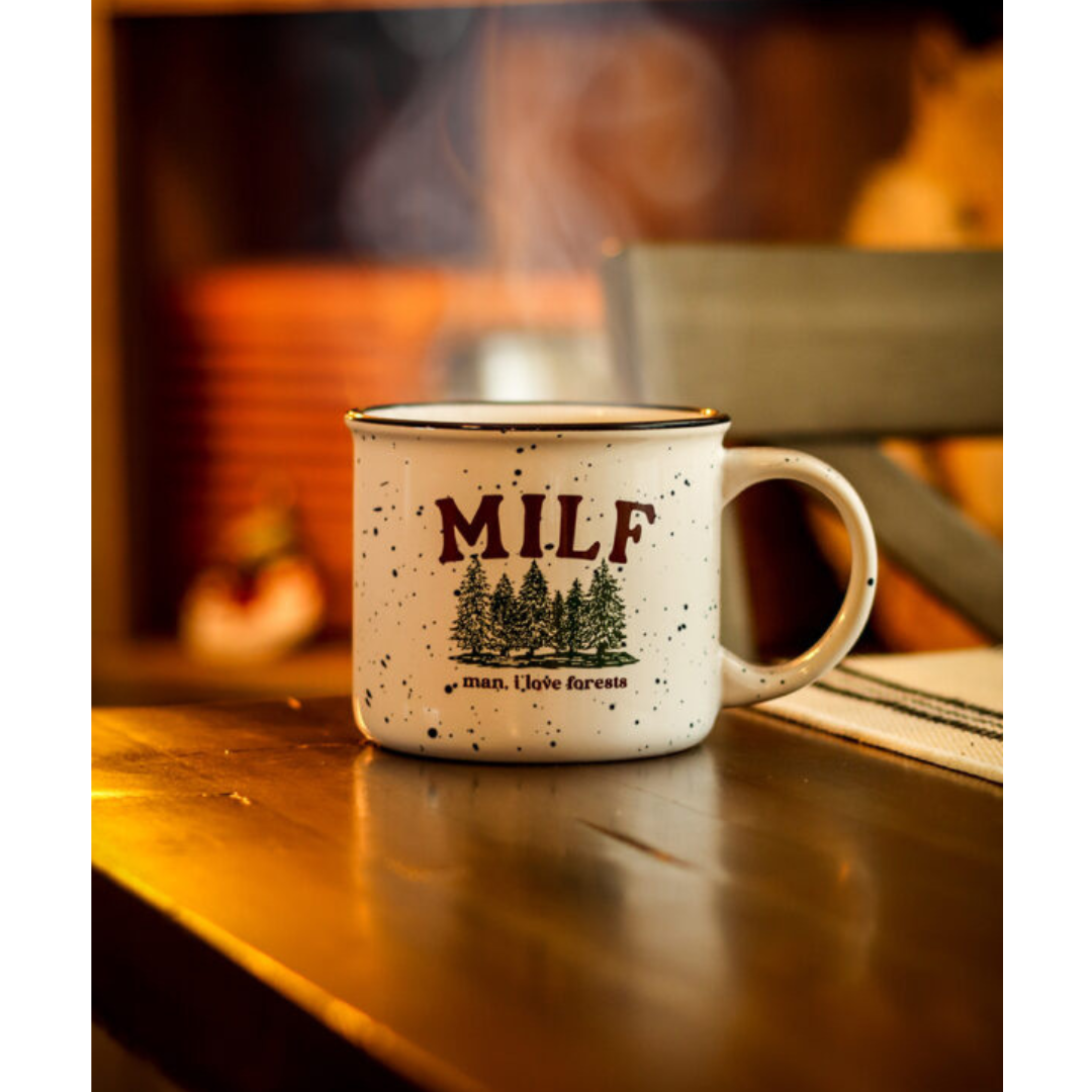 Off-white speckled ceramic mug with green forestry printed on it featuring brown lettering that reads "MILF man I love forests" sitting on brown dining table with steam coming out of the top.