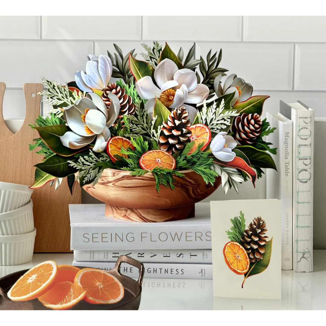 White winter magnolia bouquet pop-up greeting card resting on top of white books in white kitchen. Bouquet filled with white magnolias, pinecones, citrus fruit and winter greenery. 