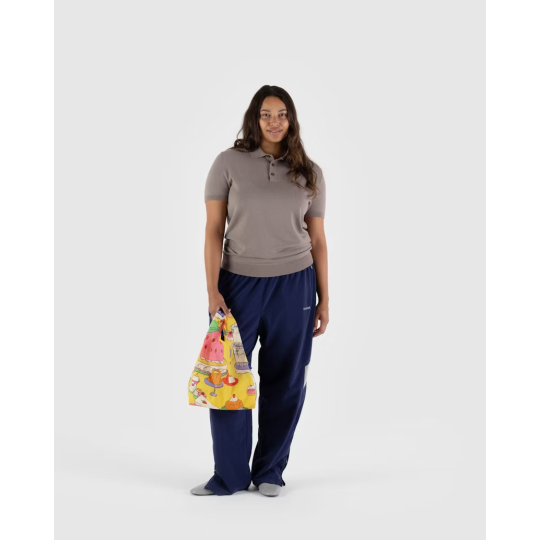 Woman carrying a yellow reusable tote bag featuring a dessert print with different types of cake all over.