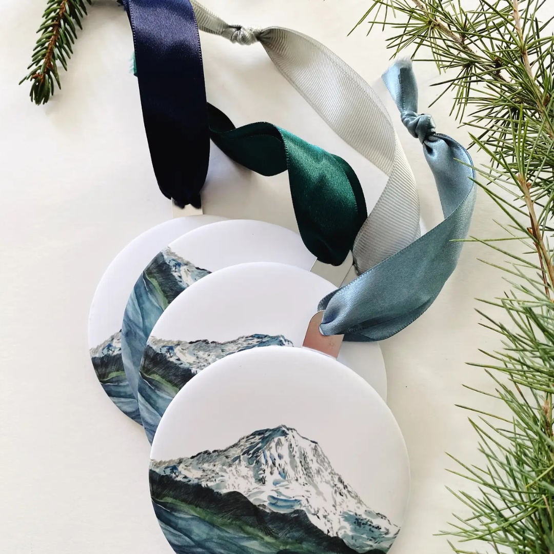 Round metal ornament featuring a watercolor painted picture of Mount Rainier in shades of blue and green. Strung by a grosgrain ribbon.