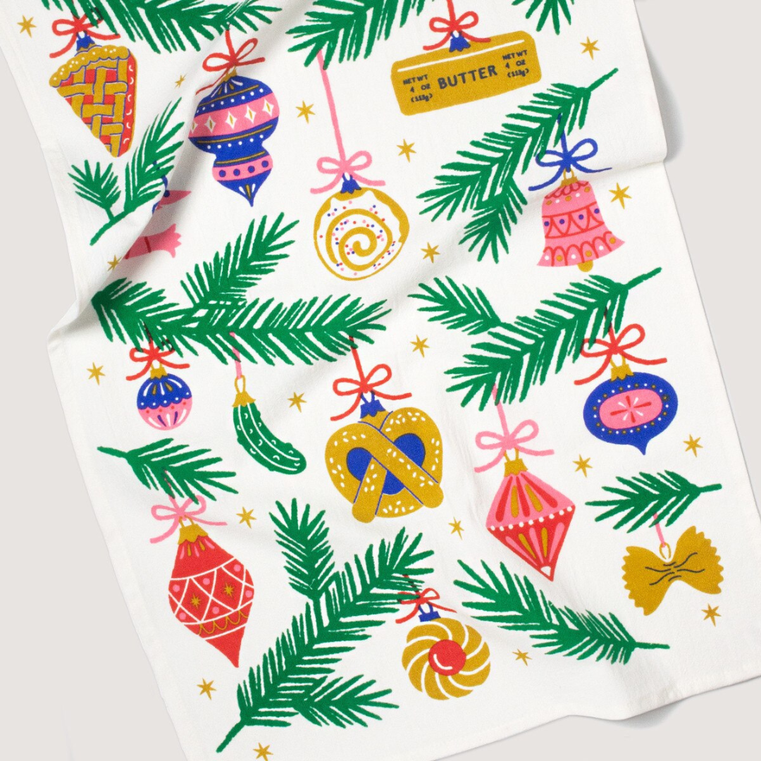 Kitchen towel with white background featuring green stems of Christmas trees and an assortment of blue, pink, red and gold Christmas ornaments.