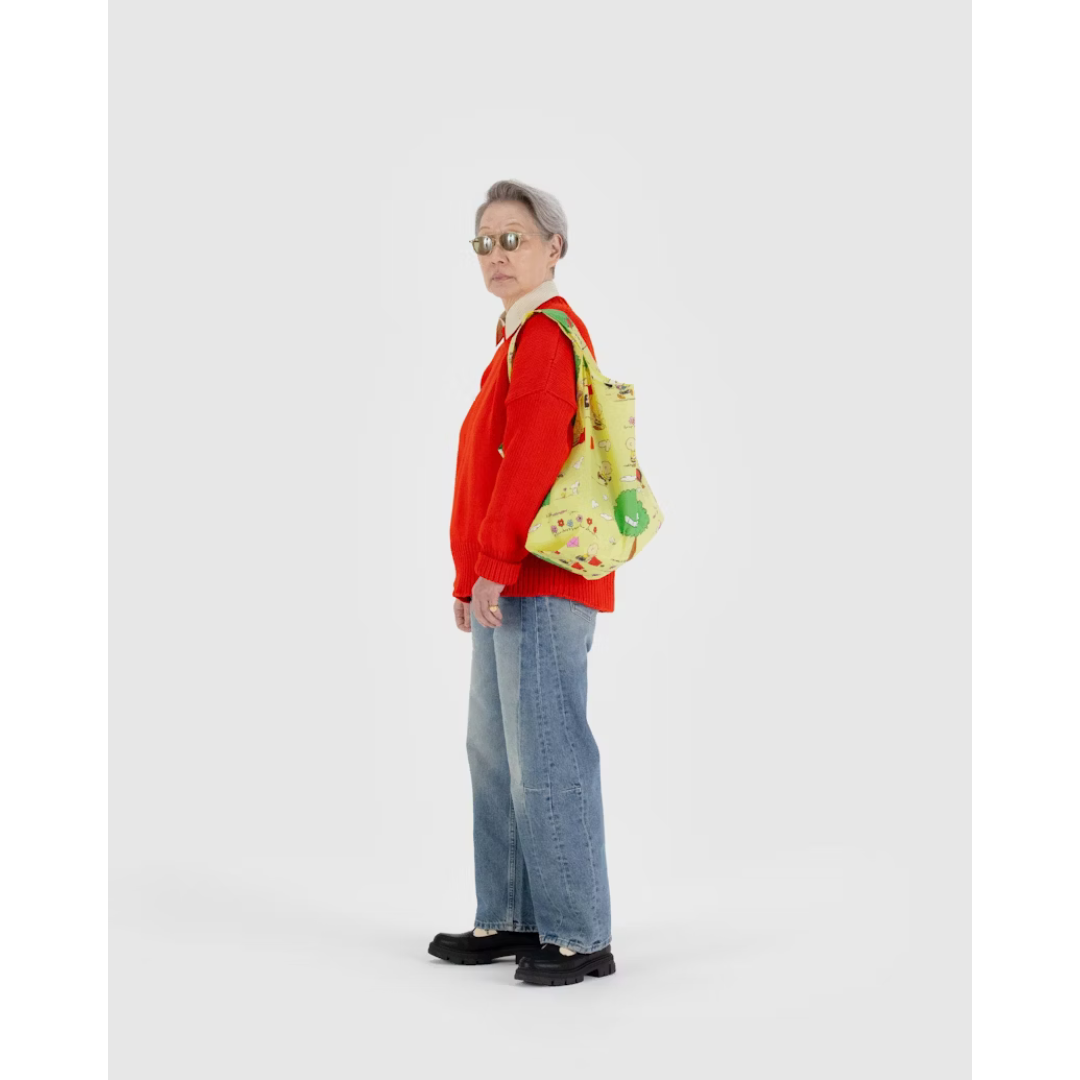 Woman holding yellowish green reusable tote bag featuring a Charlie Brown and Snoopy print flying kites into trees. 