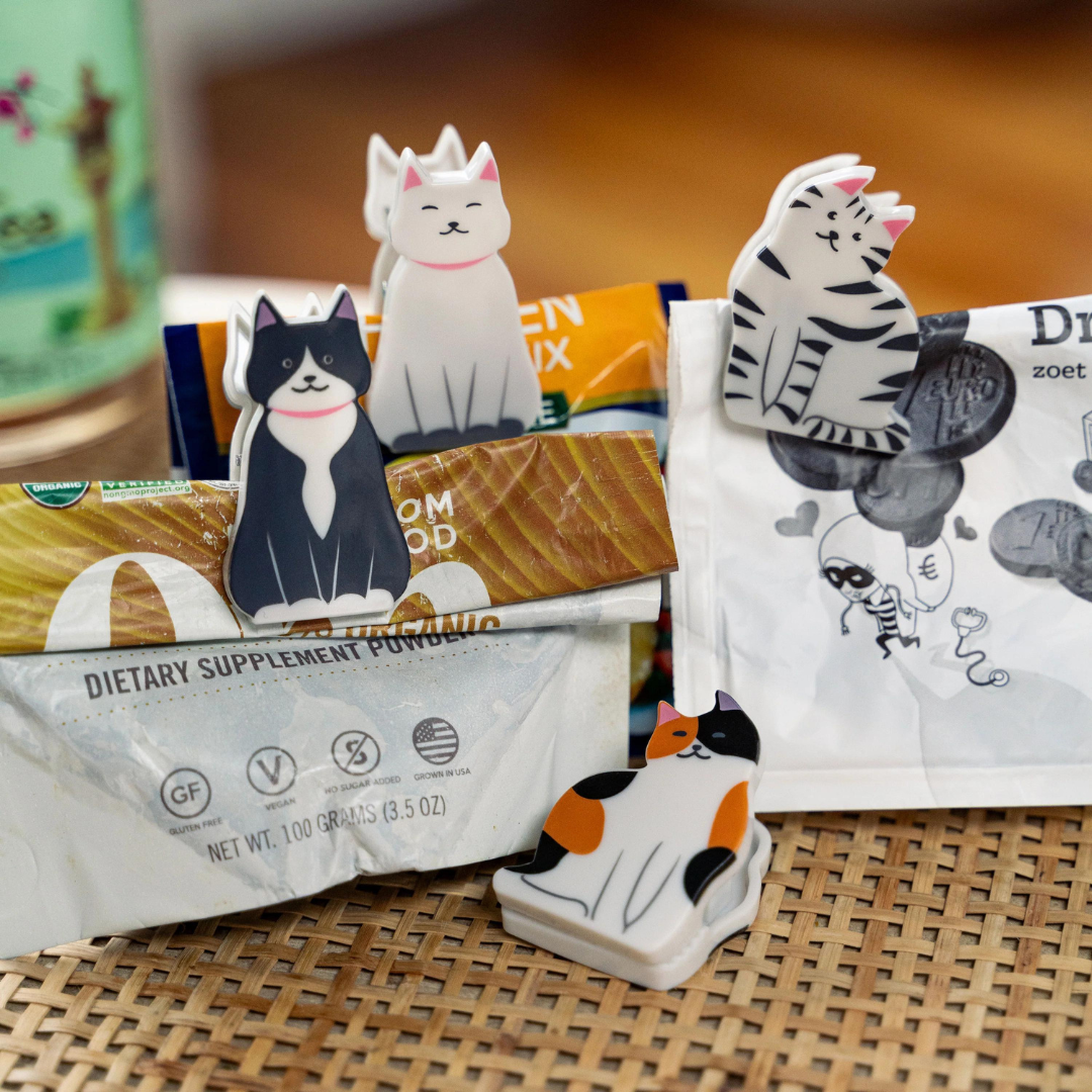 Four kitty bag clips of the black, white and orange variety attached to three chip bags, sitting on a rattan tabletop.