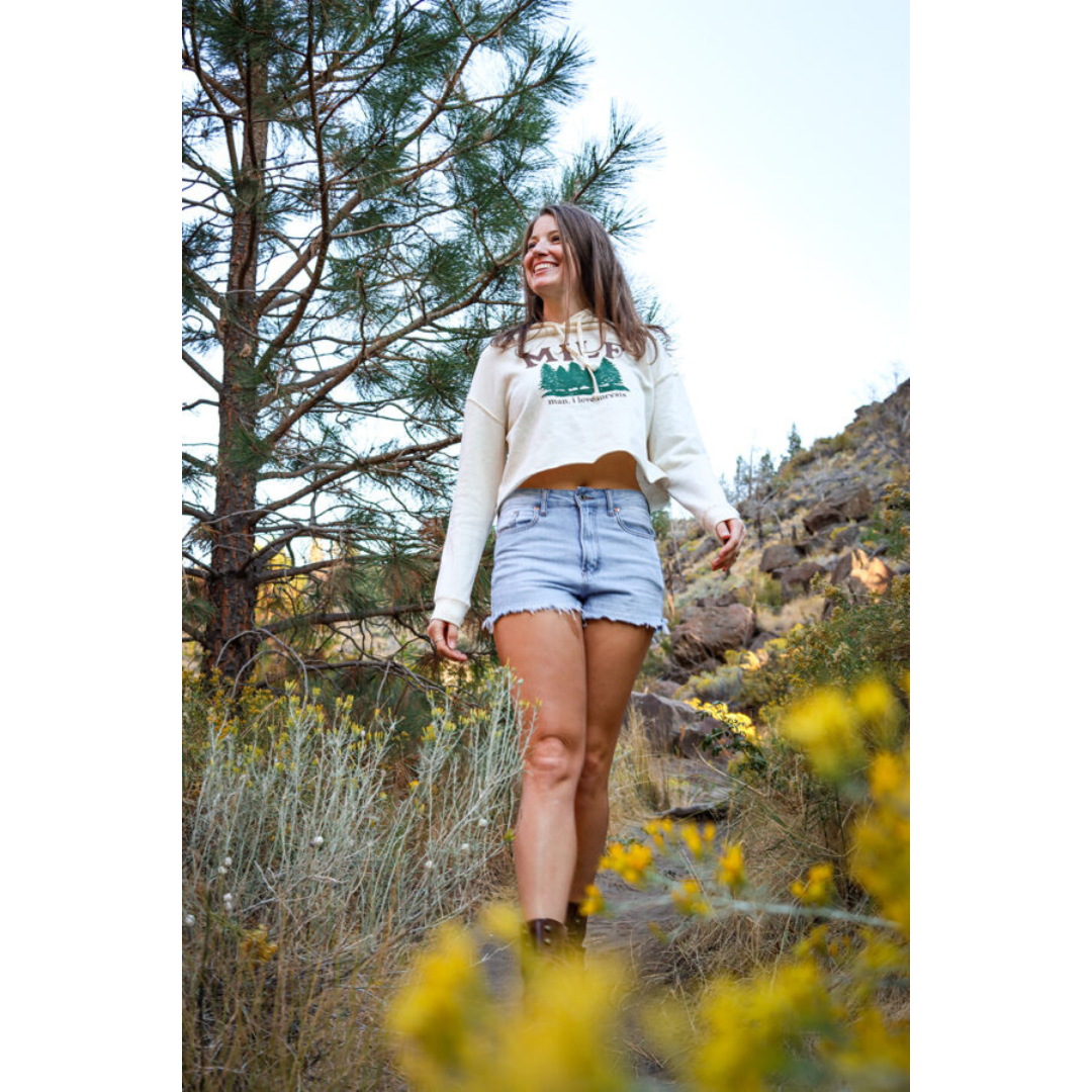 Brunette woman laughing on hiking trail wearing denim shorts and off-white hoodie with green forestry printed on it also featuring brown lettering that reads "MILF man I love forests". Yellow flowers also pictured.