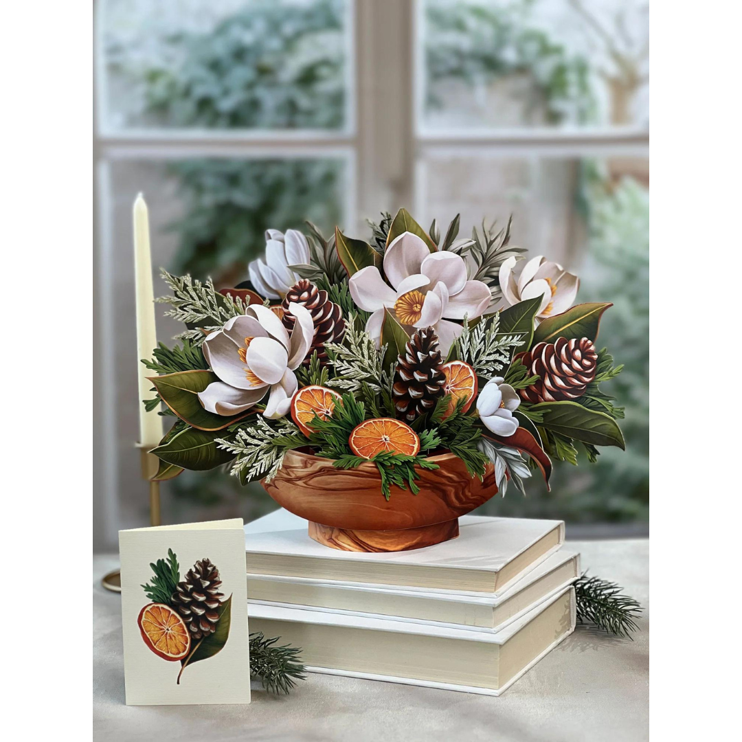 White winter magnolia bouquet pop-up greeting card resting on top of white books in front of a window. Bouquet filled with white magnolias, pinecones, citrus fruit and winter greenery. 