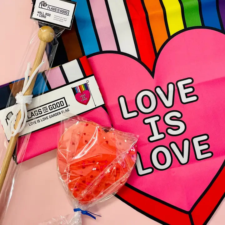 Flatlay with Love is Love flag featuring pride flag colors and and heart. Red heart lollipop lays on top of flag.