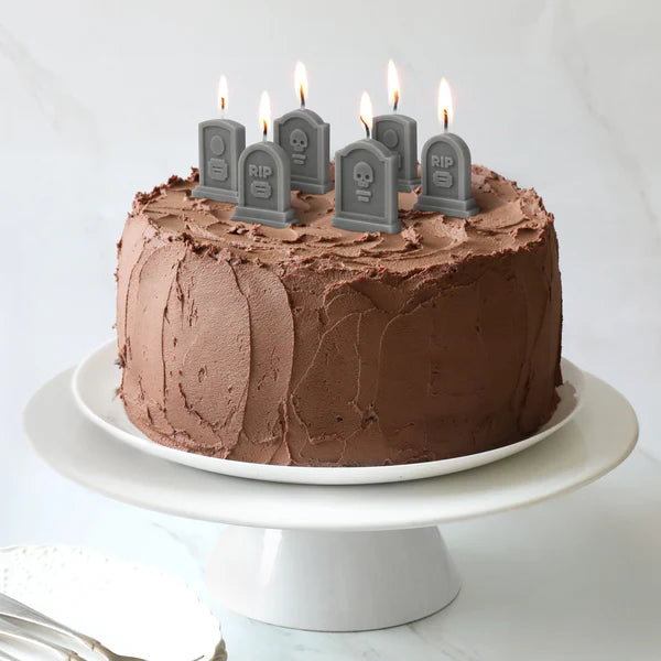 Chocolate cake with lit candles shaped like tombstones on a white cake-stand.