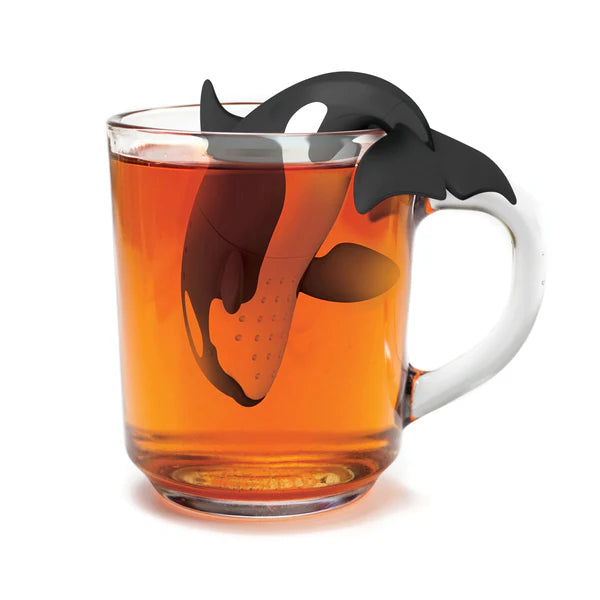 Tea infuser shaped like an orca in a glass of tea. Orca's tail hangs over the side to hold the infuser in place.