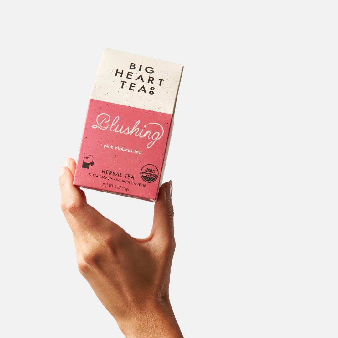 Hand holding a pink and white box of Big Heart Tea Co tea bags in the flavor "Blushing" with the words "pink hibiscus herbal tea" underneath.