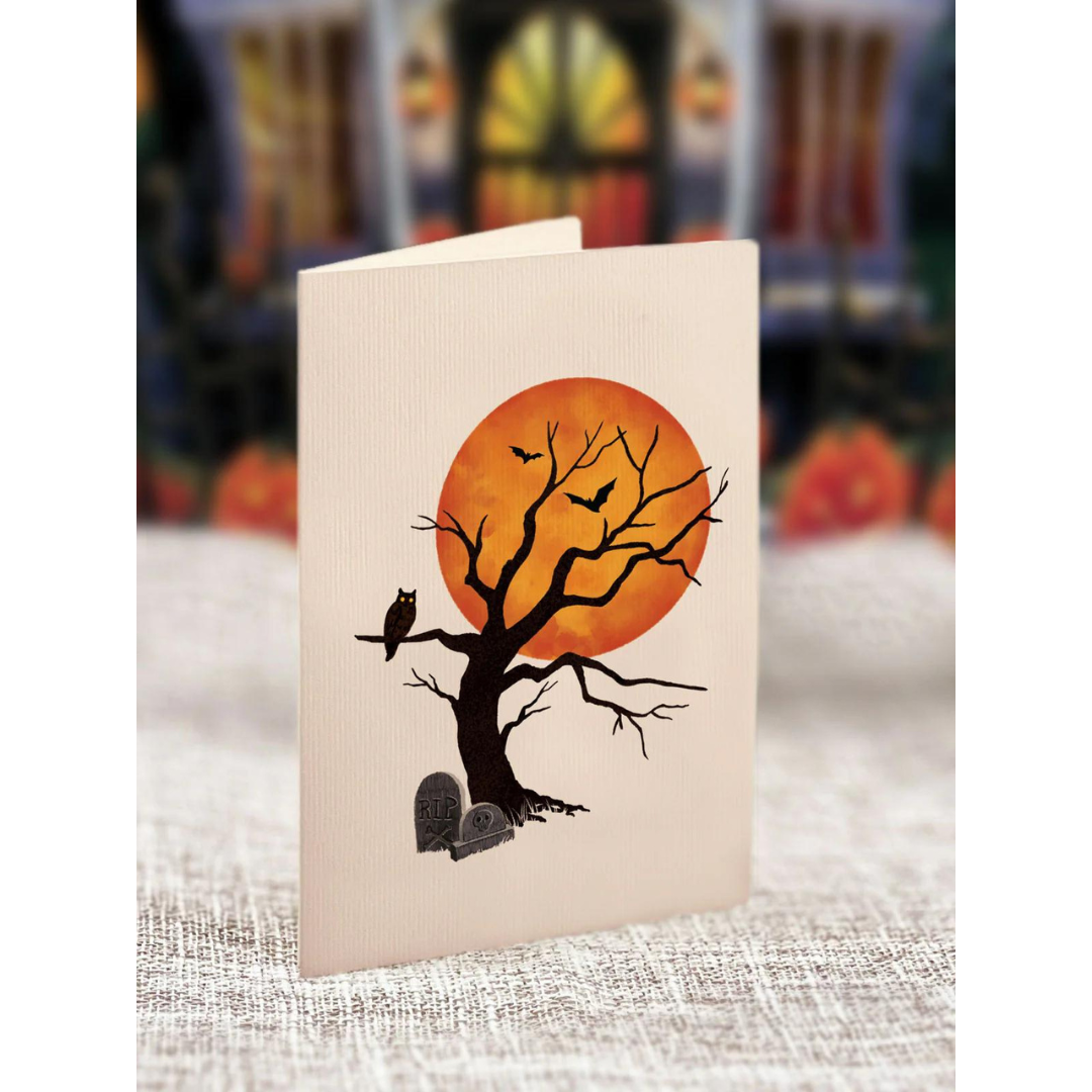 Simple white greeting card featuring a spooky tree with an owl sitting on it ad a gravestone at the foot of it, all in front of an orange moon.