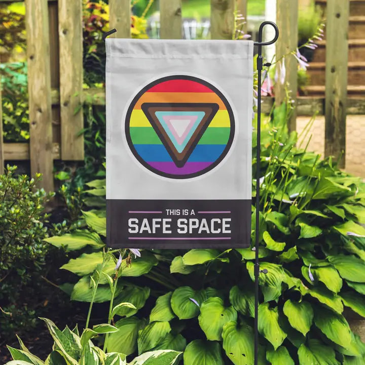 This is a safe space garden flag featuring circular inclusive pride flag in a lush green garden background.