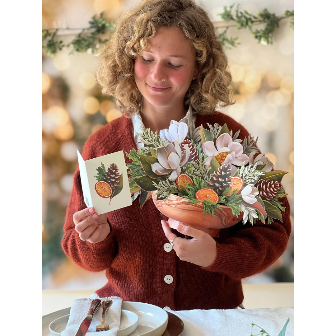 Woman in red holding greeting card and wintery floral bouquet pop-up card. Bouquet full of  white magnolias, pinecones, citrus fruit and winter greenery. 