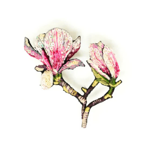 Hand embroidered and beaded magnolia brooch. Two magnolia blossoms are freshly opening on a stem, one is just a bud, the other is open. The flowers are pink and white and covered in beads and micro sequins. the stem is stitched in browns and greens.
