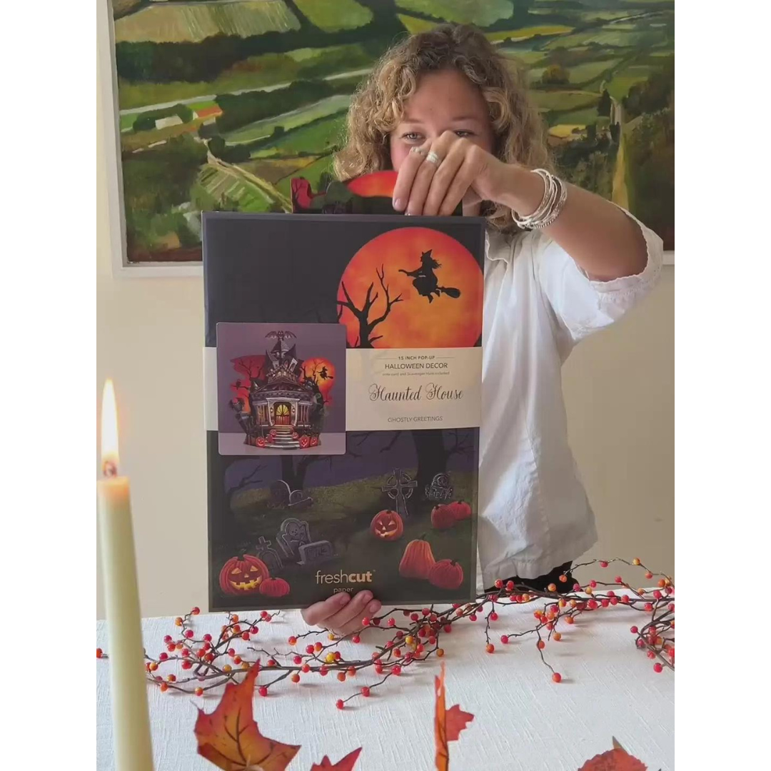 Woman in white holding packaging for Haunted House pop-up greeting card, all above an Autumn decorated table.