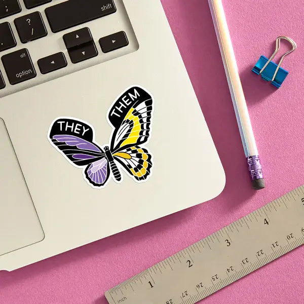 Butterfly sticker with words "they them" on wings, placed on laptop next to pencil and ruler on pink background