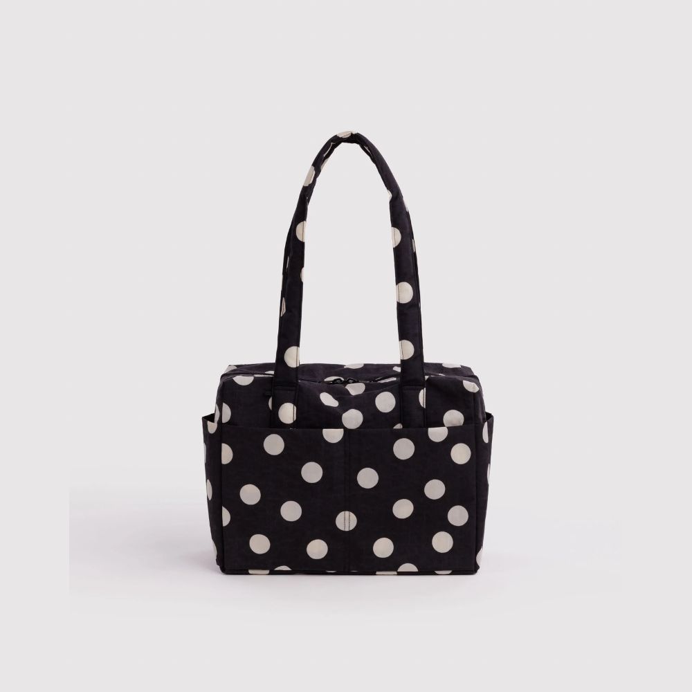 A black fabric tote bag with a white polka dot pattern, featuring two top handles and a zippered top closure, shown against a light gray background.