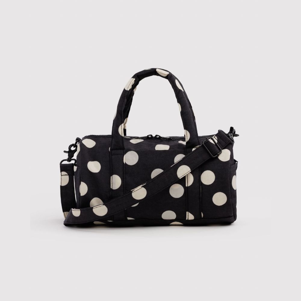 A black duffel bag with large cream polka dots, featuring dual top handles and an adjustable shoulder strap, set against a plain light gray background.