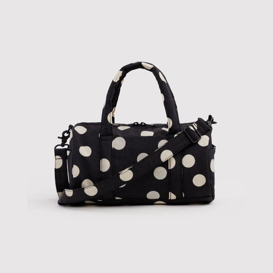 A black duffel bag with large cream polka dots, featuring dual top handles and an adjustable shoulder strap, set against a plain light gray background.