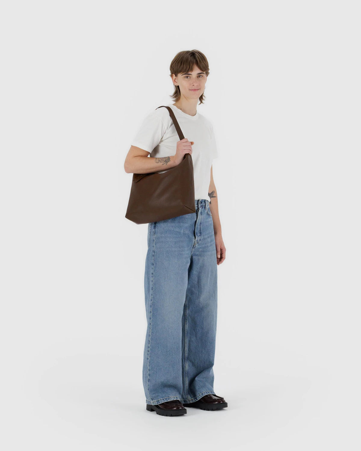 Person wearing a white t-shirt, blue jeans, and holding a brown leather bag on a white background